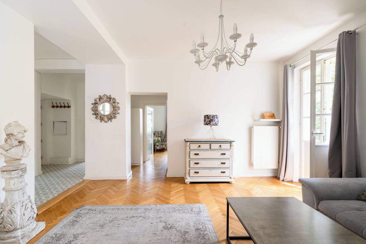 Uma sala de estar ampla com um candelabro, um sofá, uma mesa de centro e um aparador. Há um espelho na parede e uma estátua no chão.