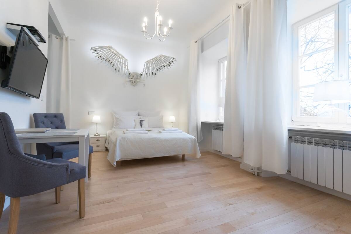 Quarto com cama, escrivaninha, cadeira, televisão e janela com cortinas.