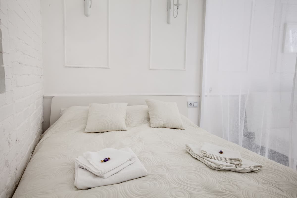 Uma cama bem arrumada com almofadas e toalhas dobradas, uma parede de tijolos brancos e uma janela com cortinas leves.