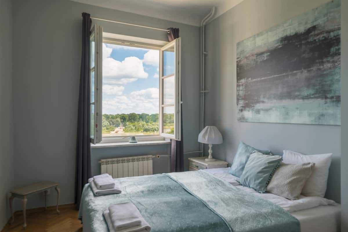 Quarto com cama, mesa de cabeceira, luminária, janela com cortinas e um quadro na parede.