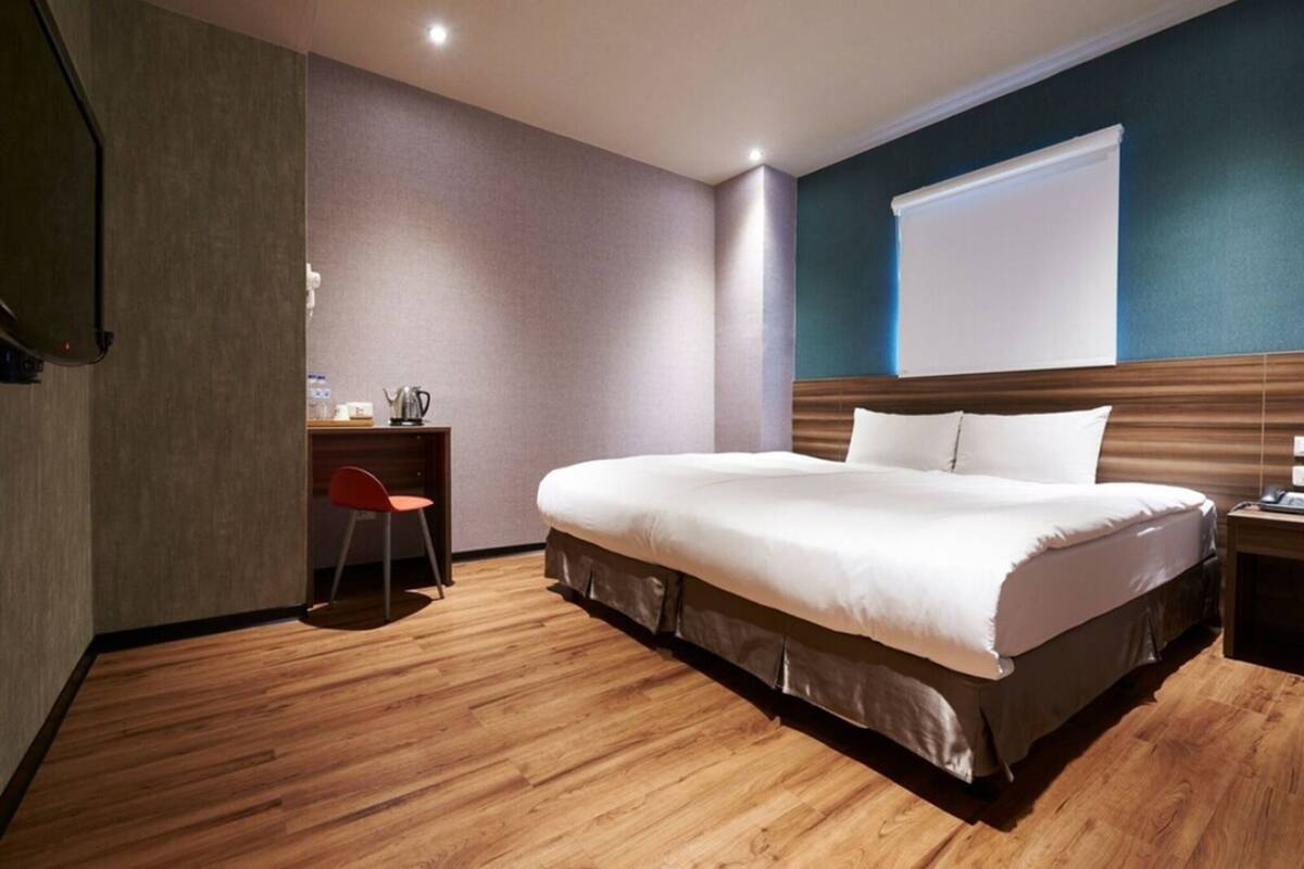 A modern hotel room with a large bed, a desk with a chair, and a window with blinds.