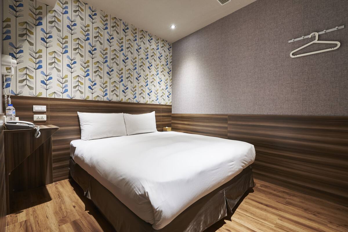 A hotel room with a large bed, wooden headboard, a patterned wallpaper, a grey textured wall, and a white hanger on the wall.