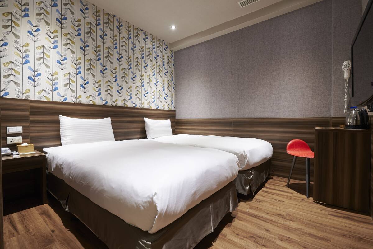 A hotel room with two beds, a wooden headboard, a desk with a chair, and a patterned wallpaper.