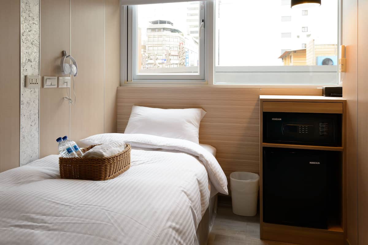 A modern hotel room with a bed, a small desk with a microwave, a window with a view of buildings, and a hanging light fixture.