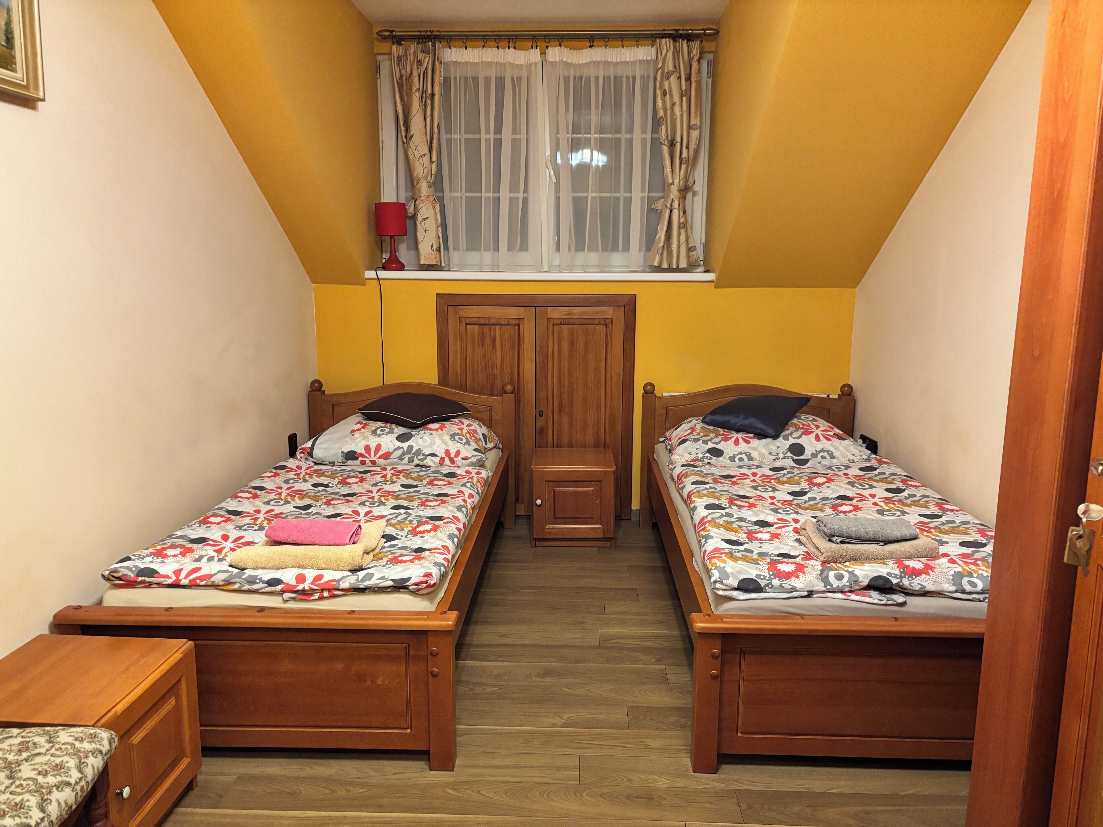 A small, cozy bedroom with two wooden beds, a nightstand, a wooden wardrobe, and a window with curtains.