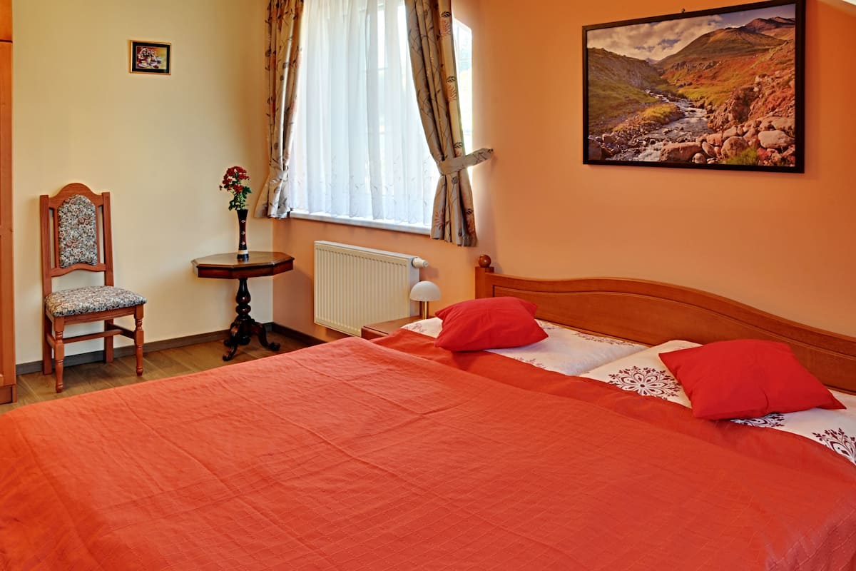 A bedroom with a large bed, a chair, a small table, a radiator, and a window with curtains.