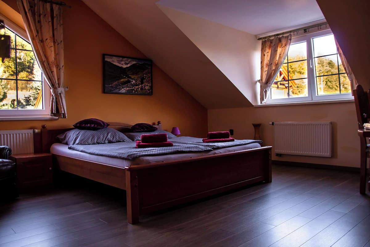 A bedroom with a sloped ceiling, a bed with pillows and a blanket, a nightstand, a lamp, and a window with curtains.