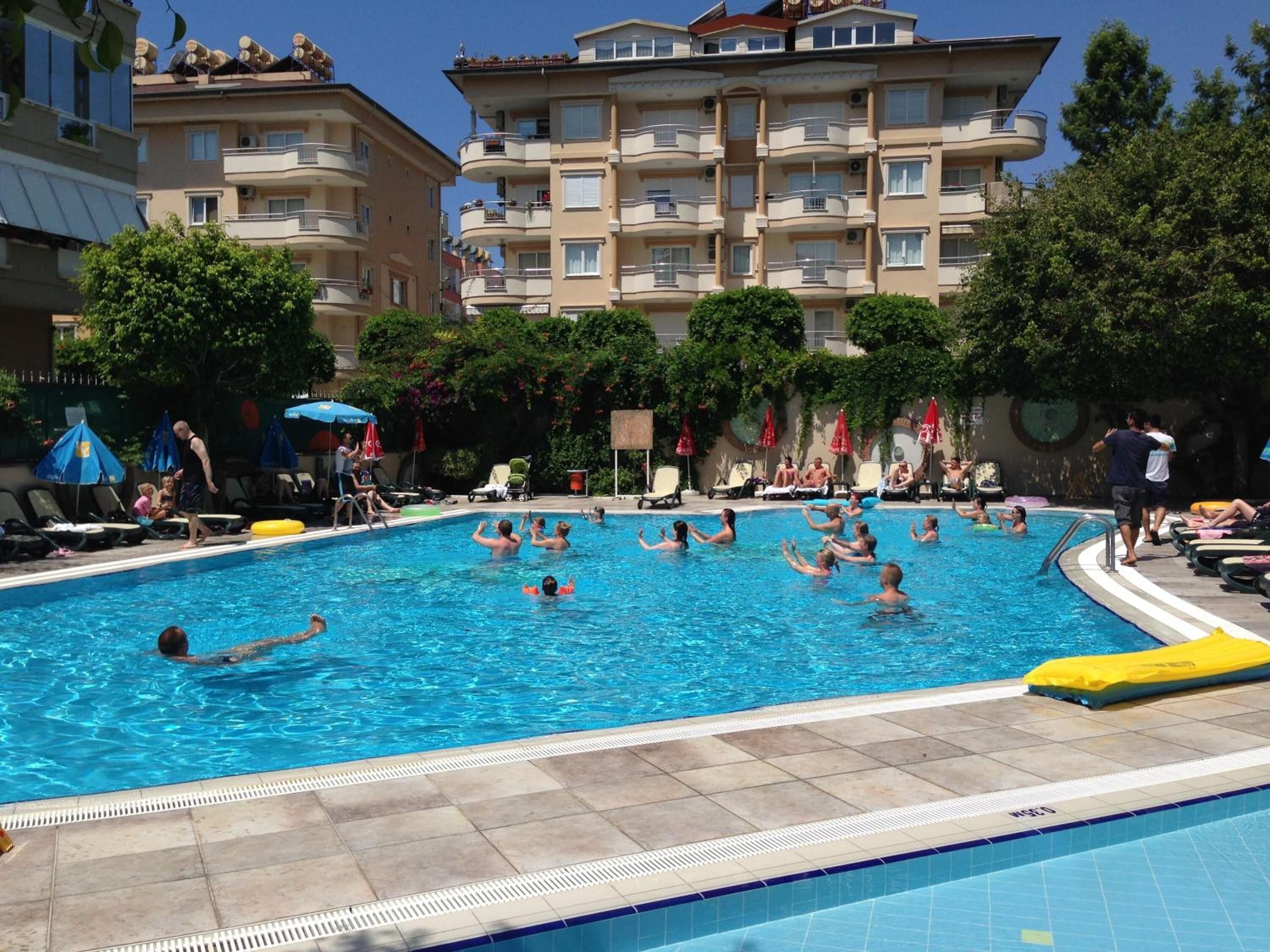 Una piscina rodeada de sillas de descanso y sombrillas, con un edificio de varios pisos al fondo.