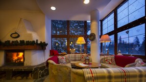 A cozy living room with a fireplace, comfortable seating, and large windows with a view of snow-covered trees. - Penzion Pleso (Štrba)
