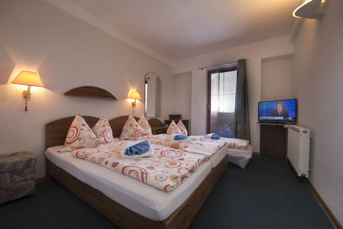 A hotel room with a double bed, two bedside tables, a TV, and a window with curtains.