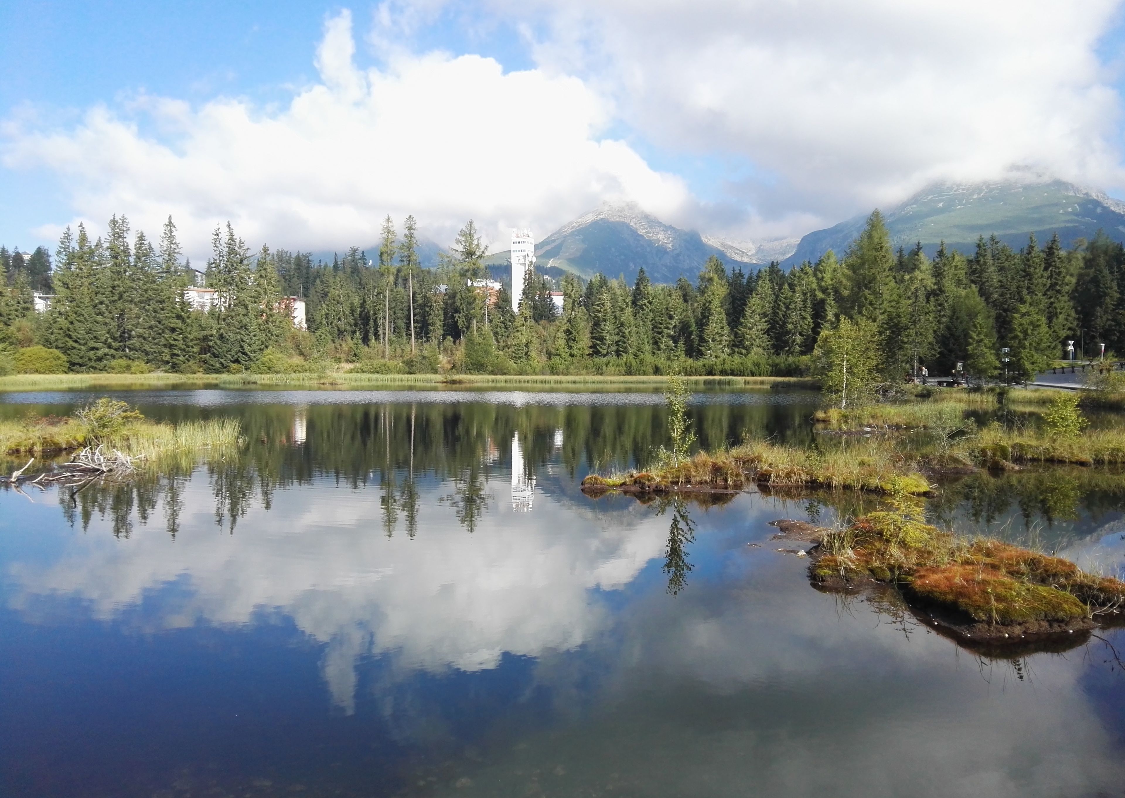 Un lago tranquilo con reflejos de árboles y montañas, rodeado de un bosque denso y un pequeño edificio al fondo.