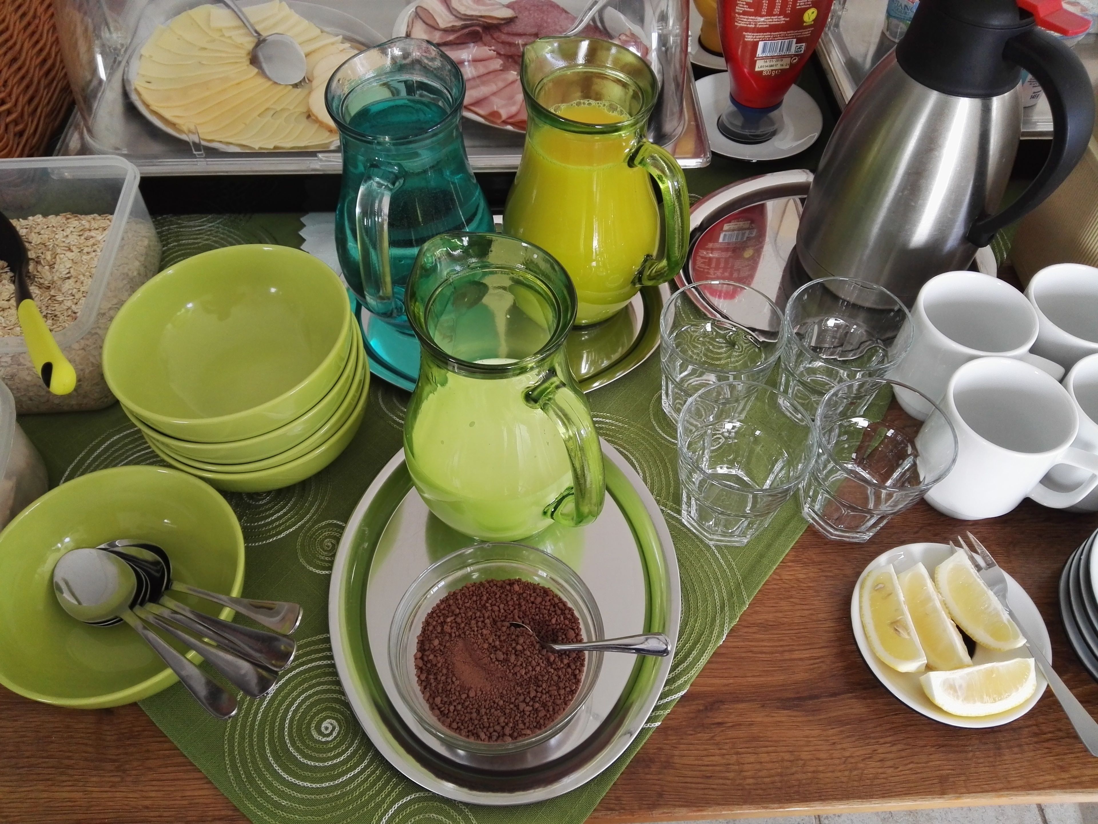 A breakfast table with various food items and drinks, including a green pitcher, a glass of orange juice, and a plate of sliced fruit.