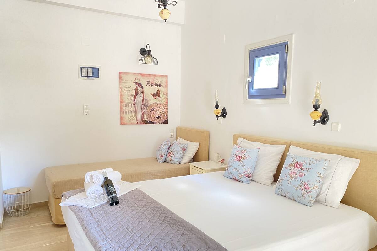 A hotel room with a large bed, two smaller beds, floral pillows, wall art, and a small side table.