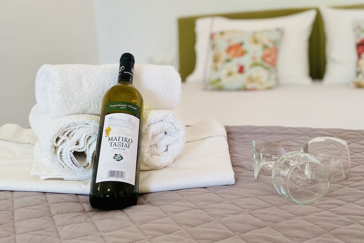 A bed with a quilted cover, a bottle of wine, a rolled towel, and a glass on a bedside table.