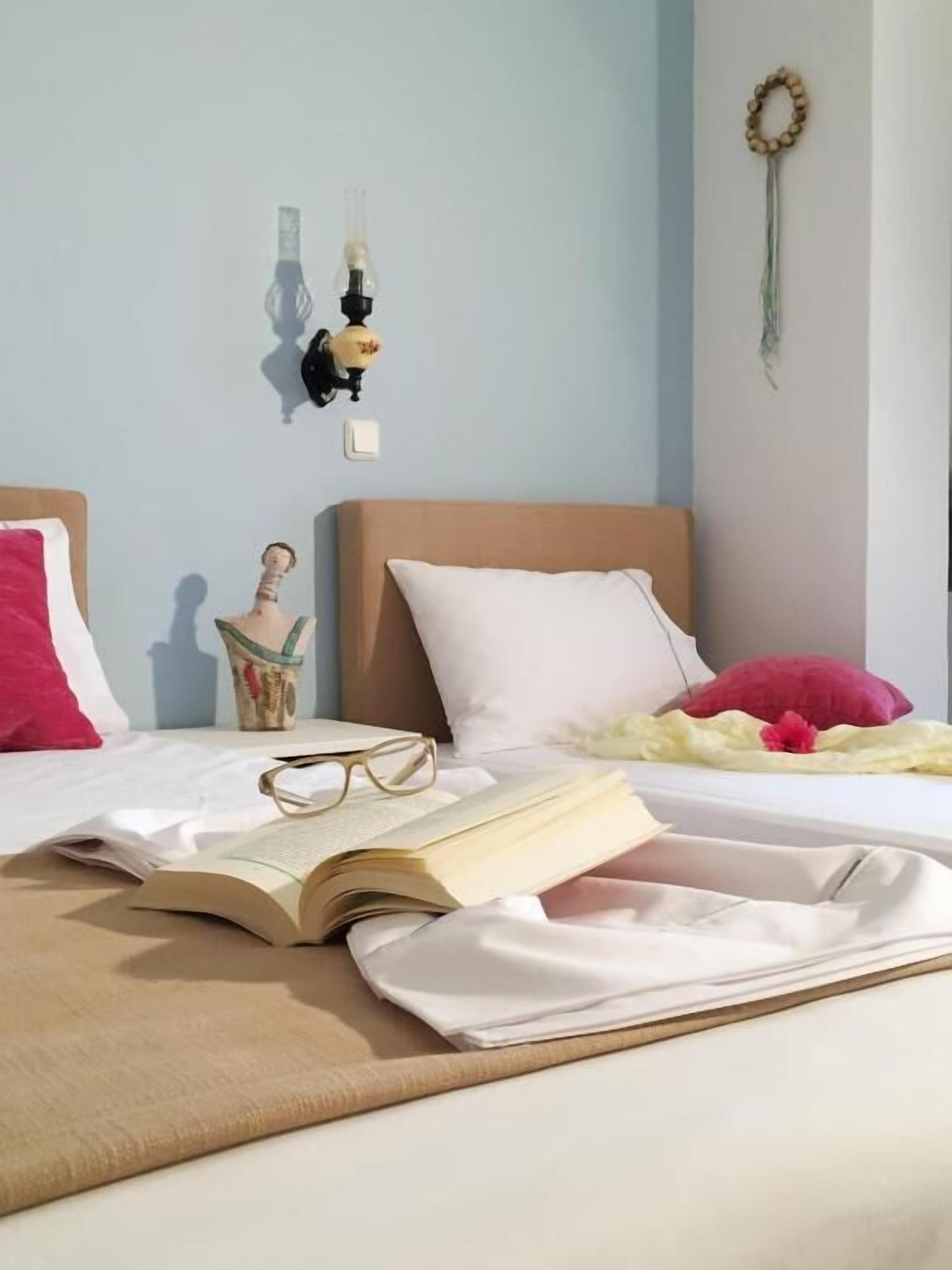 Una cama bien hecha con un libro, gafas y un adorno encima. Hay una lámpara de pared y un atrapasueños en la pared.