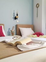 Una cama bien hecha con un libro, gafas y un adorno encima. Hay una lámpara de pared y un atrapasueños en la pared.