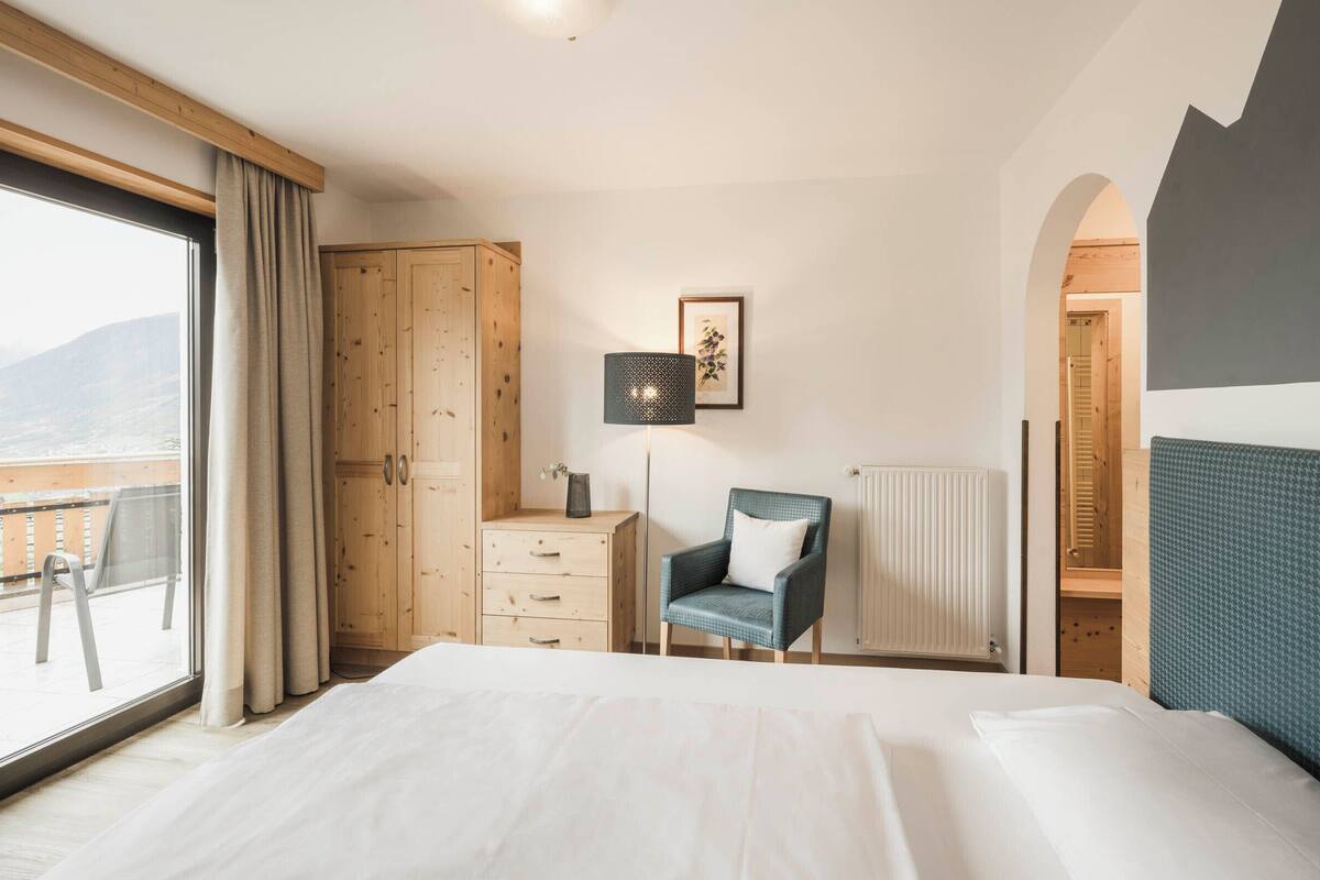 A bedroom with a large bed, a wooden wardrobe, a desk with a lamp, a chair, and a view of the outdoors.