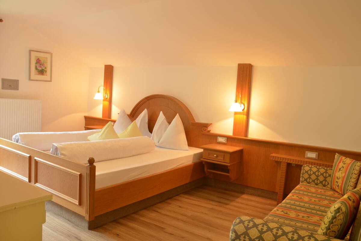 A hotel room with a wooden bed, bedside tables, a nightstand, a sofa, and wall-mounted lamps.