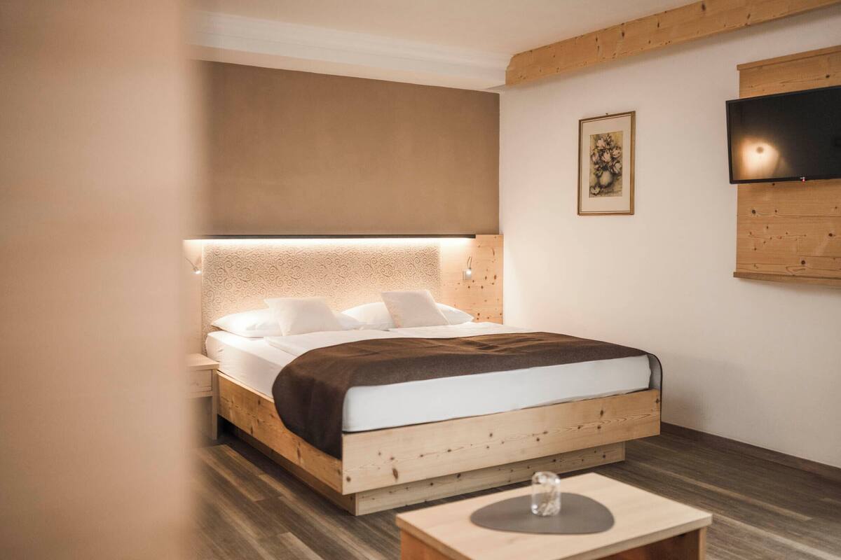 A modern bedroom with a wooden bed, a bedside table, a flat-screen TV mounted on the wall, and a framed artwork above the bed.