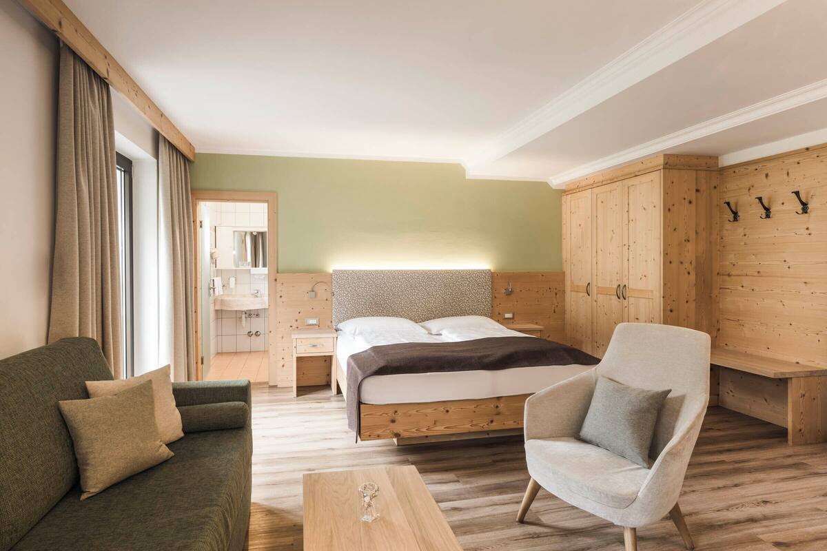 A hotel room with a bed, a sofa, a chair, a wooden wardrobe, and a bathroom visible through an open door.