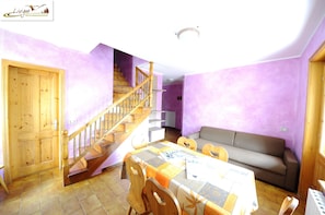 Una habitación con una escalera de madera, un sofá, una mesa de comedor con sillas y una puerta de madera.