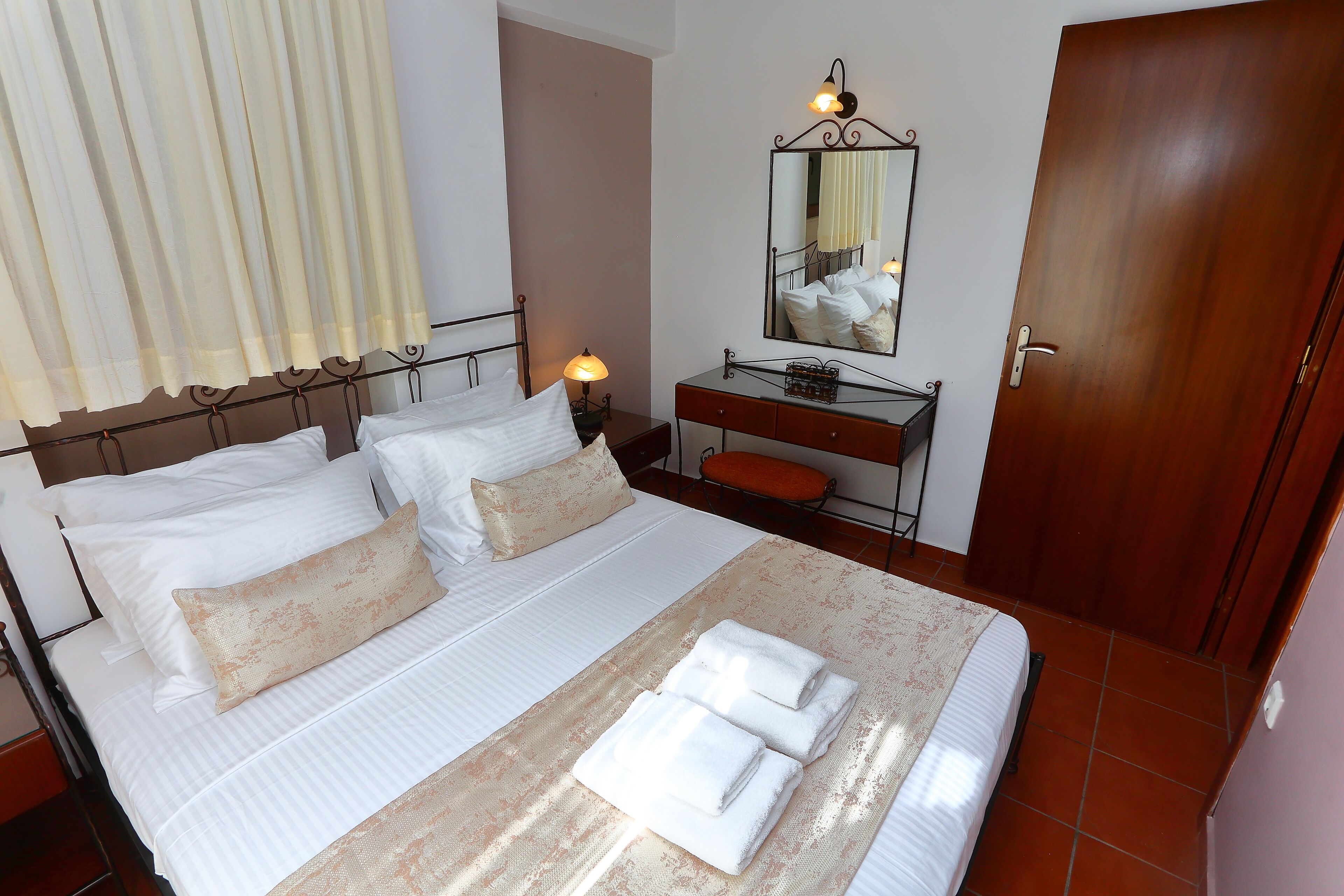 Un letto rifatto con lenzuola bianche e copriletto color beige, un comodino con una lampada, uno specchio sopra il tavolo e una porta in legno.