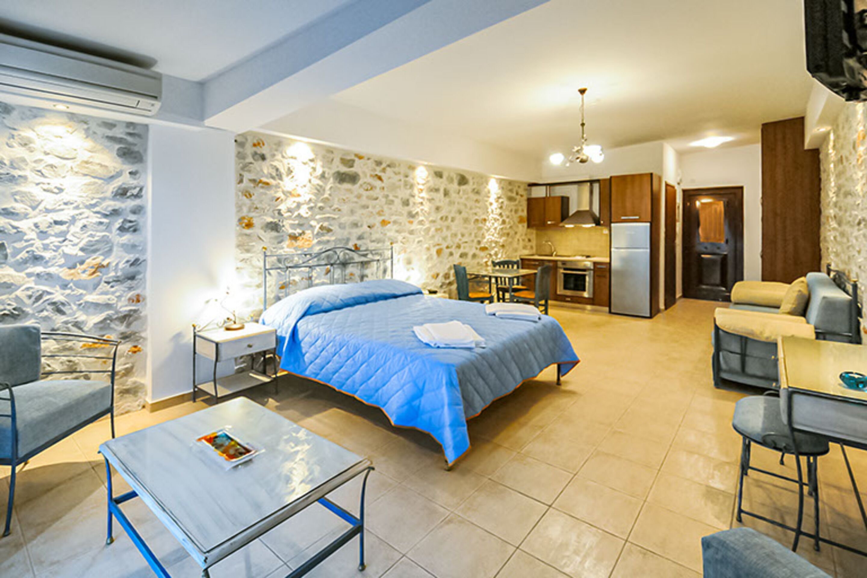 A compact room with a bed, a small table, a chair, a kitchenette, and a sofa.