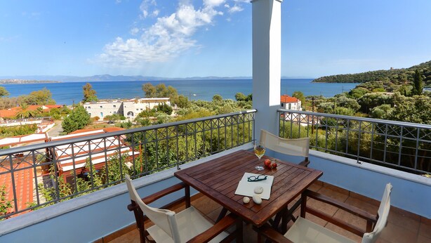 A balcony with a wooden table, chairs, and a view of the sea and distant buildings. - Vathi Hotel (East Mani)