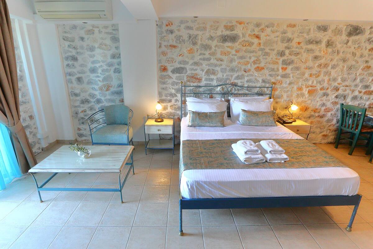 A bedroom with a stone wall, a bed with white linens, a small table with a lamp, and a chair.