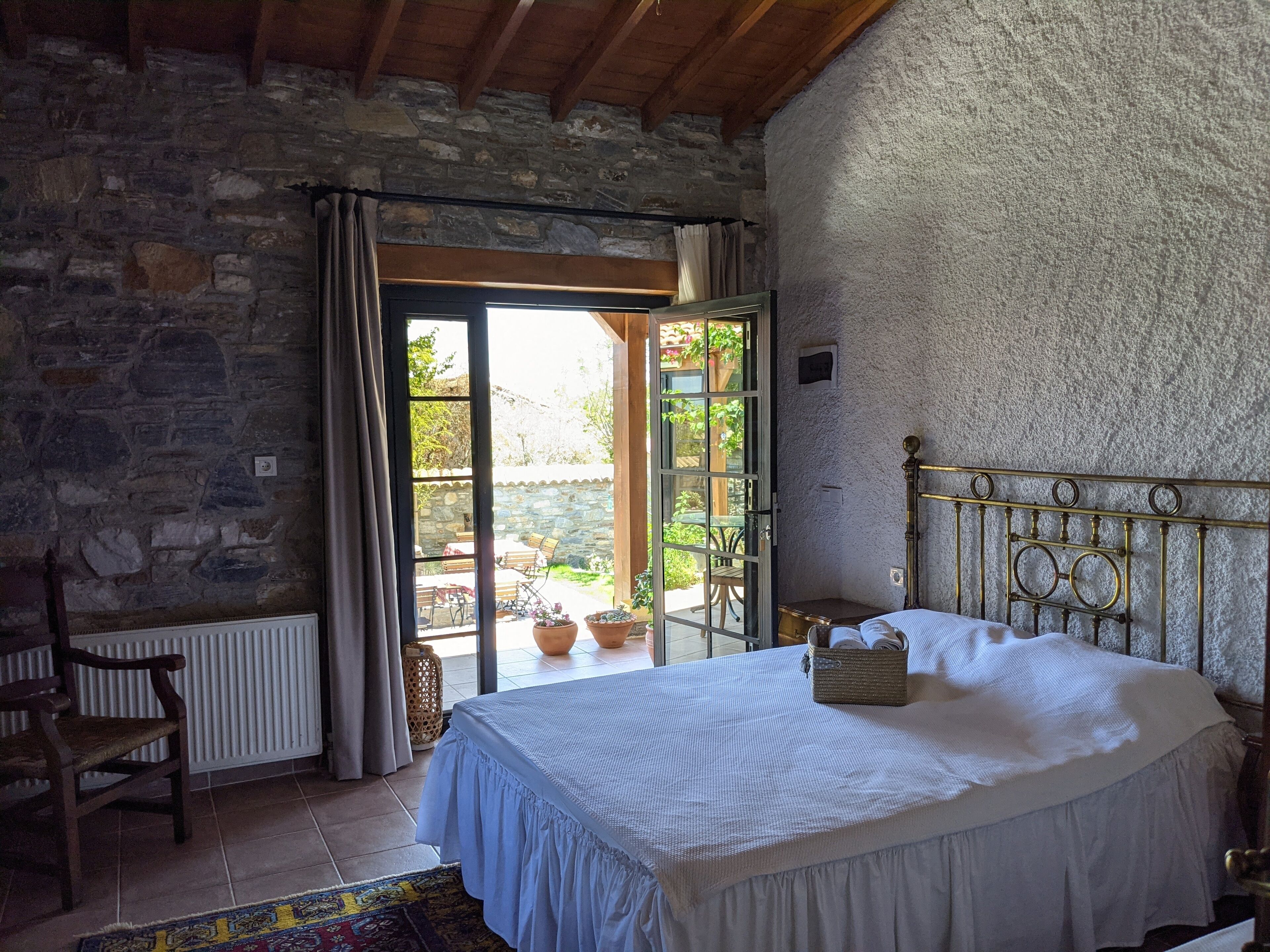 Um quarto com parede de pedra, cama com colcha branca, cadeira de madeira e vista do jardim através de uma porta de vidro.