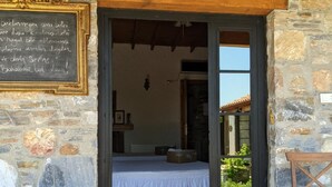 A stone-walled room with a bed, a wooden door, and a chalkboard with text. There are potted plants outside. - Bahab Guest House (Soke)