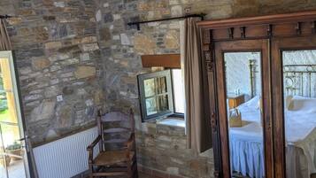 A rustic room with a stone wall, a wooden ceiling, a bed with a white bedspread, a wooden chair, a mirror, and a window with curtains.