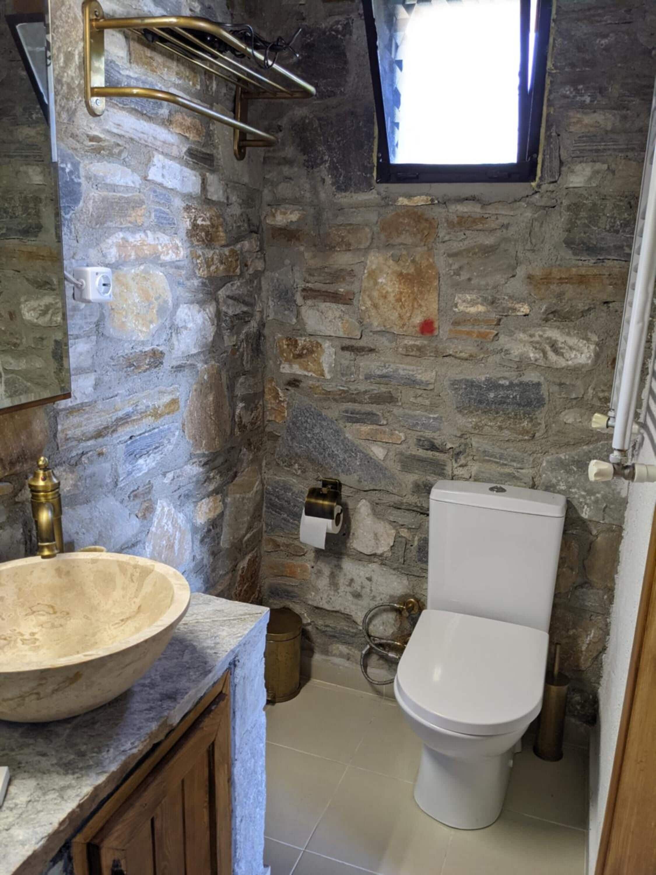 A bathroom with a stone wall, a wooden cabinet, a marble sink, a toilet, and a window with a view.