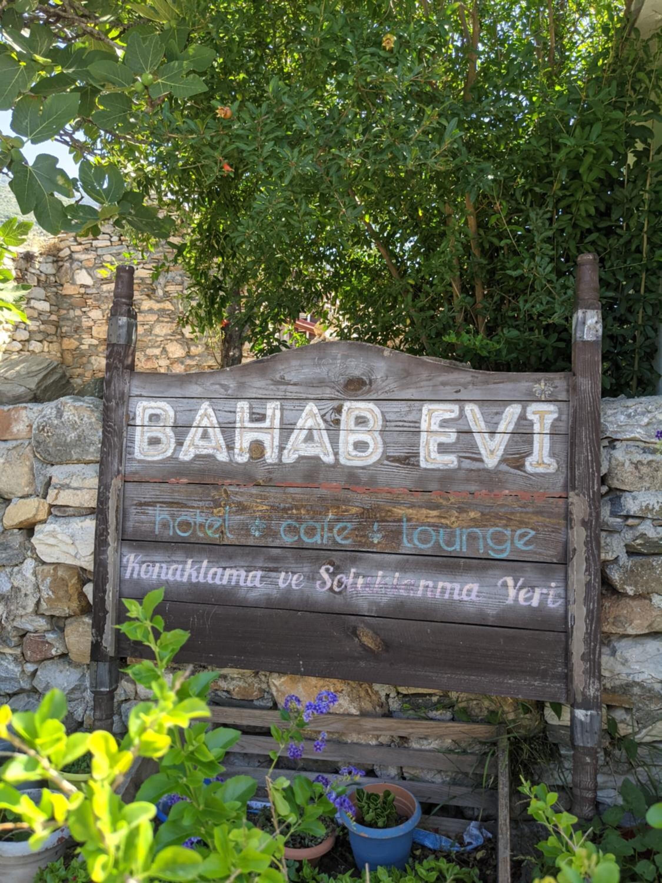 Une enseigne en bois sur laquelle on peut lire « BAHAB EVI » et « hôtel, café, salon », placée devant un mur de pierre avec de la verdure en arrière-plan.