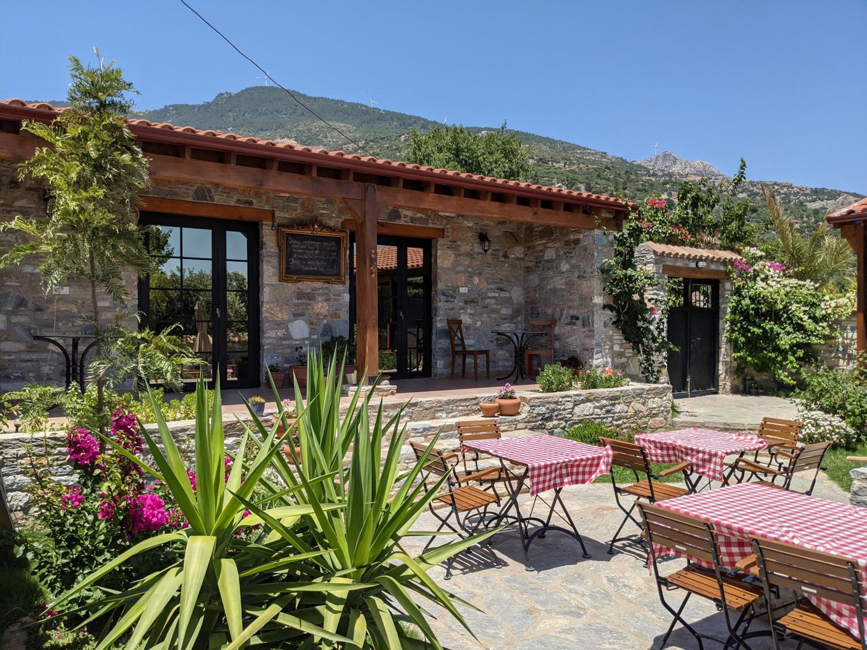A stone building with a red-tiled roof, outdoor seating with checkered tablecloths, and potted plants.