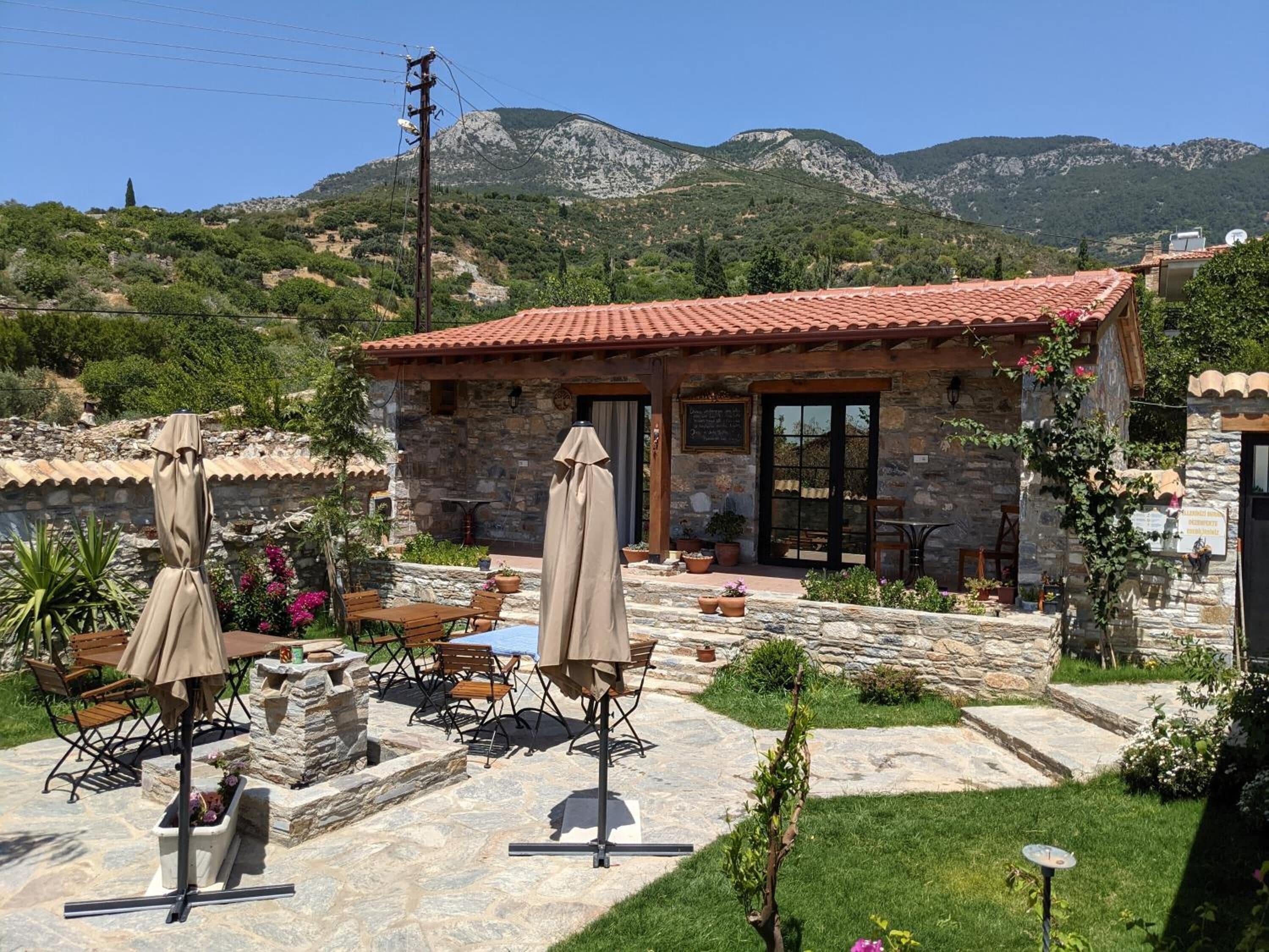 A stone building with a red-tiled roof, outdoor seating, and a view of mountains.