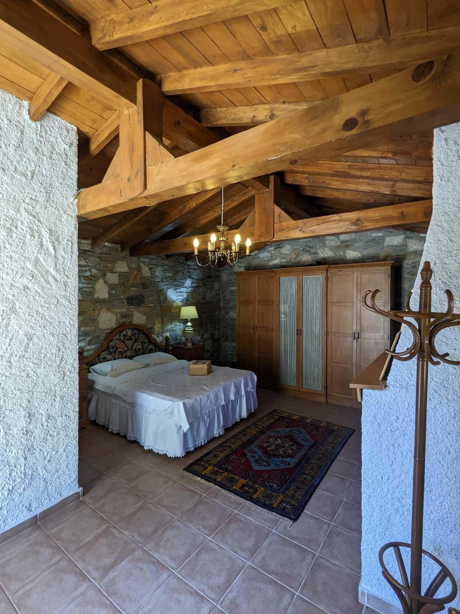 A bedroom with a stone wall, wooden ceiling beams, a bed with white linens, a nightstand, a lamp, a wardrobe, and a patterned rug.