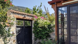 A stone building with a black gate, a stone pathway leading to a wooden pergola, and a garden with various plants and flowers. - Bahab Guest House (Soke)