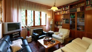 A living room with a television, leather sofas, a coffee table with decorative items, a bookshelf, and a window with curtains. - B&B Villa Sans Souci (Civitavecchia)