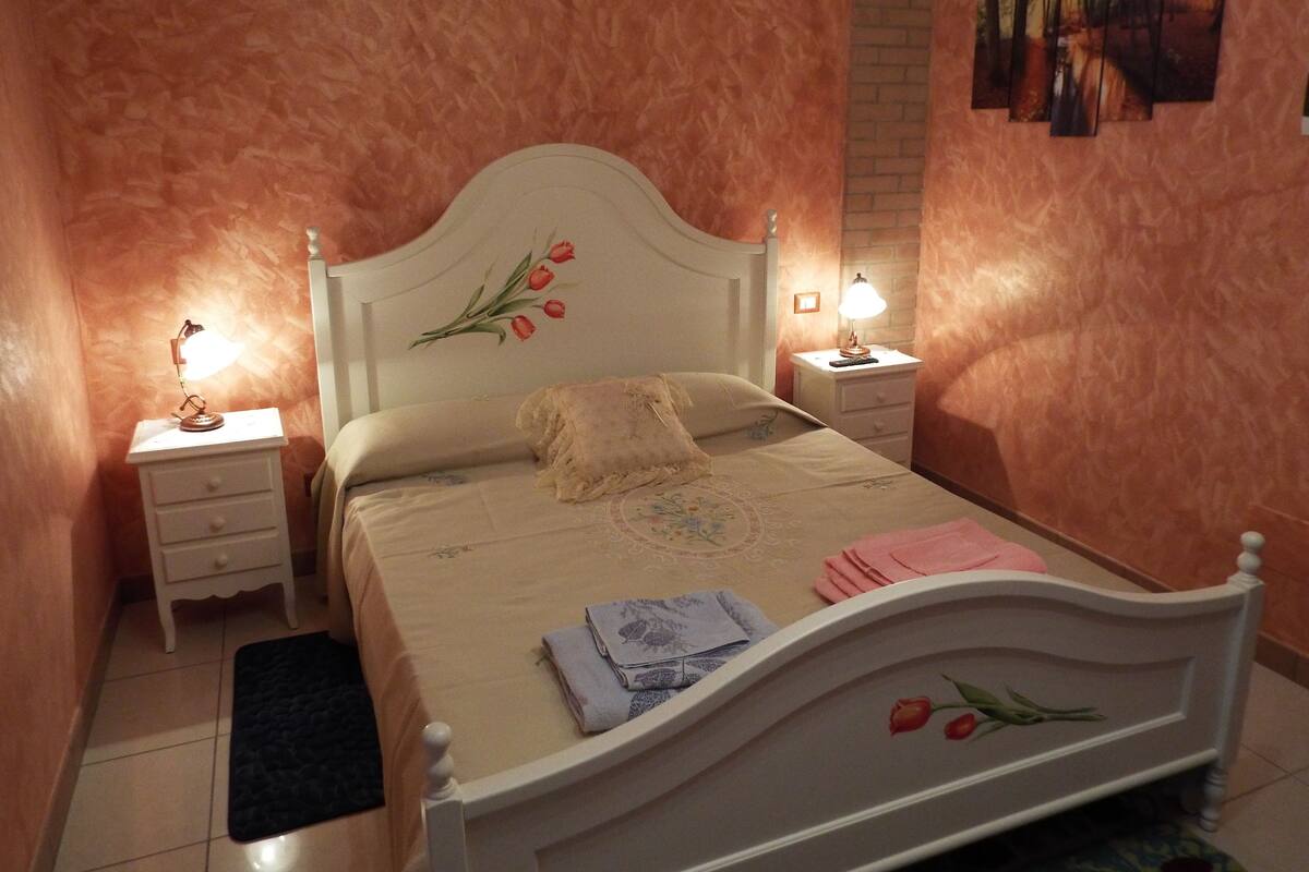 A bedroom with a bed, two bedside tables, a nightstand, a rug, and a wall with artwork.
