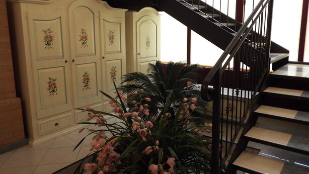 Una escalera con barandilla negra, una pared blanca con diseños florales y una planta grande en maceta con flores rosadas.