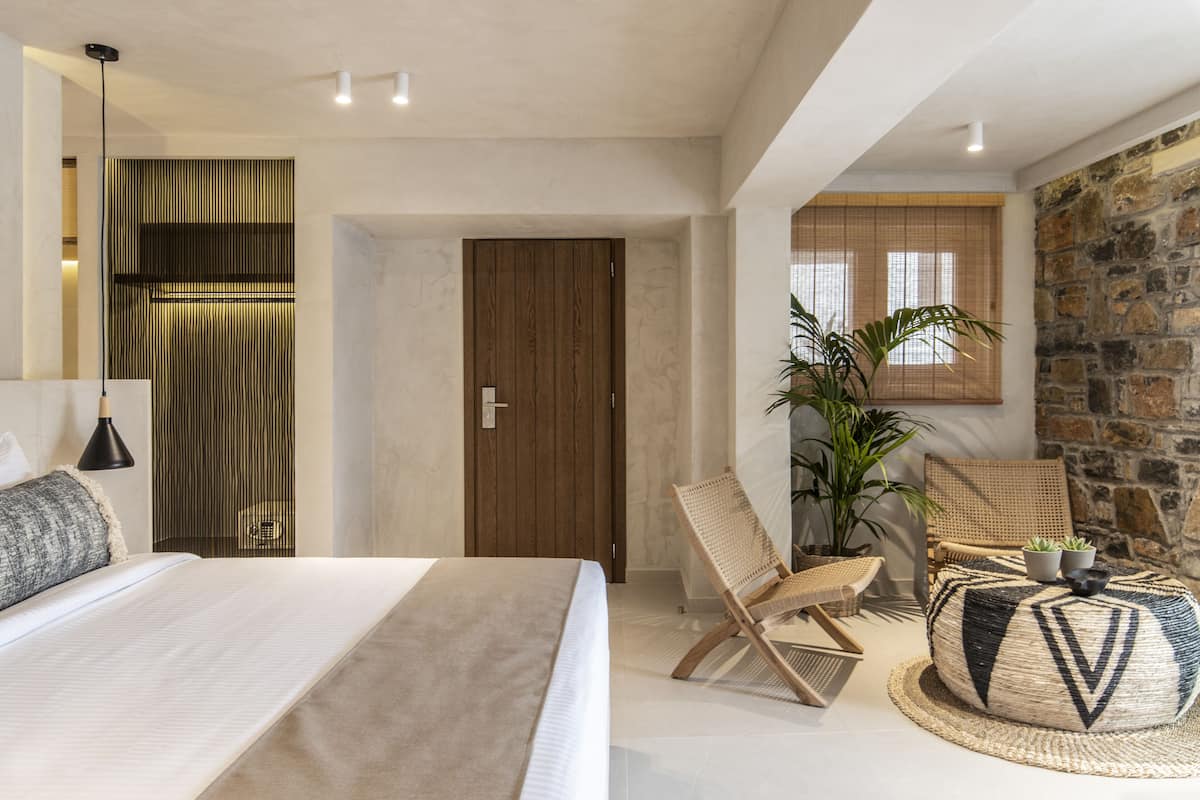 A modern bedroom with a large bed, a wooden door, a stone wall, and a wicker chair.