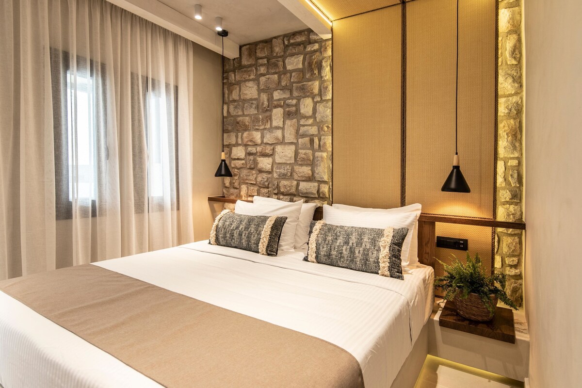 A neatly made bed with white linens and two decorative pillows, a stone accent wall, and a small bedside table with a plant.