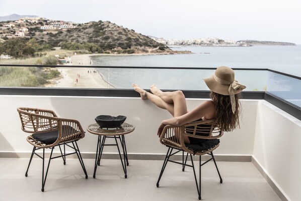 Ein Balkon mit Korbstühlen und einem kleinen Tisch, der Blick auf einen Strand und eine hügelige Landschaft bietet.