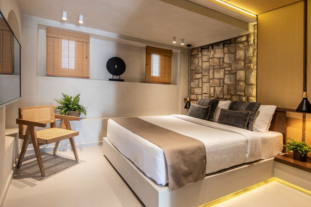 A modern bedroom with a large bed, a wooden chair, a small table, and a stone accent wall.