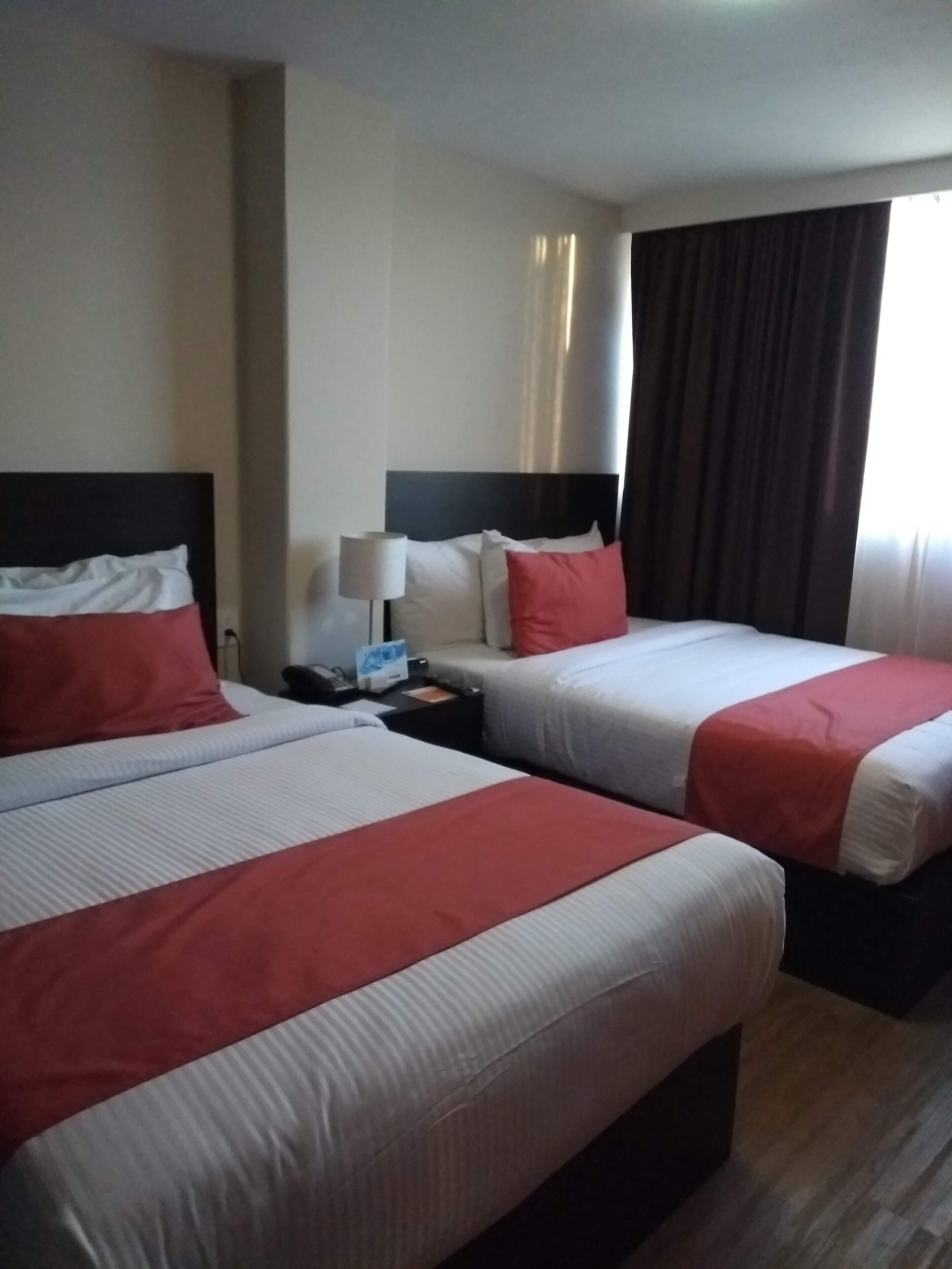 Hotellihuone, jossa on kaksi sänkyä, ja molemmissa on valkoiset ja punaiset vuodevaatteet. Huoneessa on myös yöpöytä, jossa on lamppu, sekä suuri ikkuna, jossa on verhot.