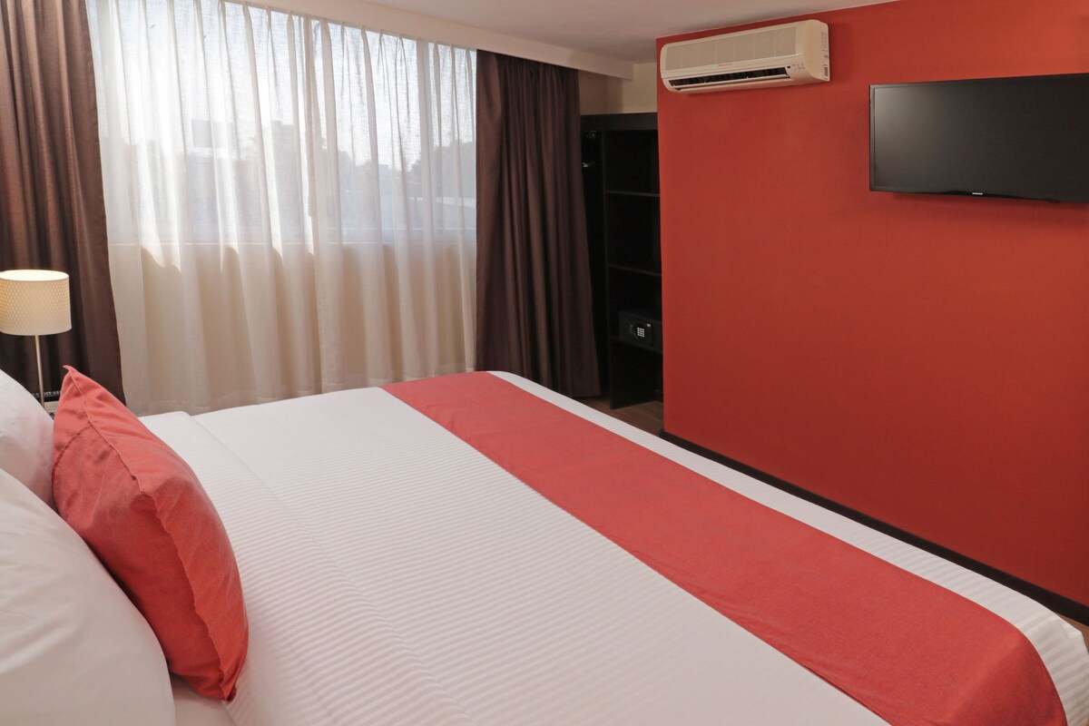A hotel room with a bed, a TV mounted on the wall, and an air conditioning unit.