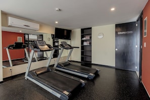 Sala de ginástica com esteiras, televisão, relógio e uma porta com a inscrição "GIMNASIO".