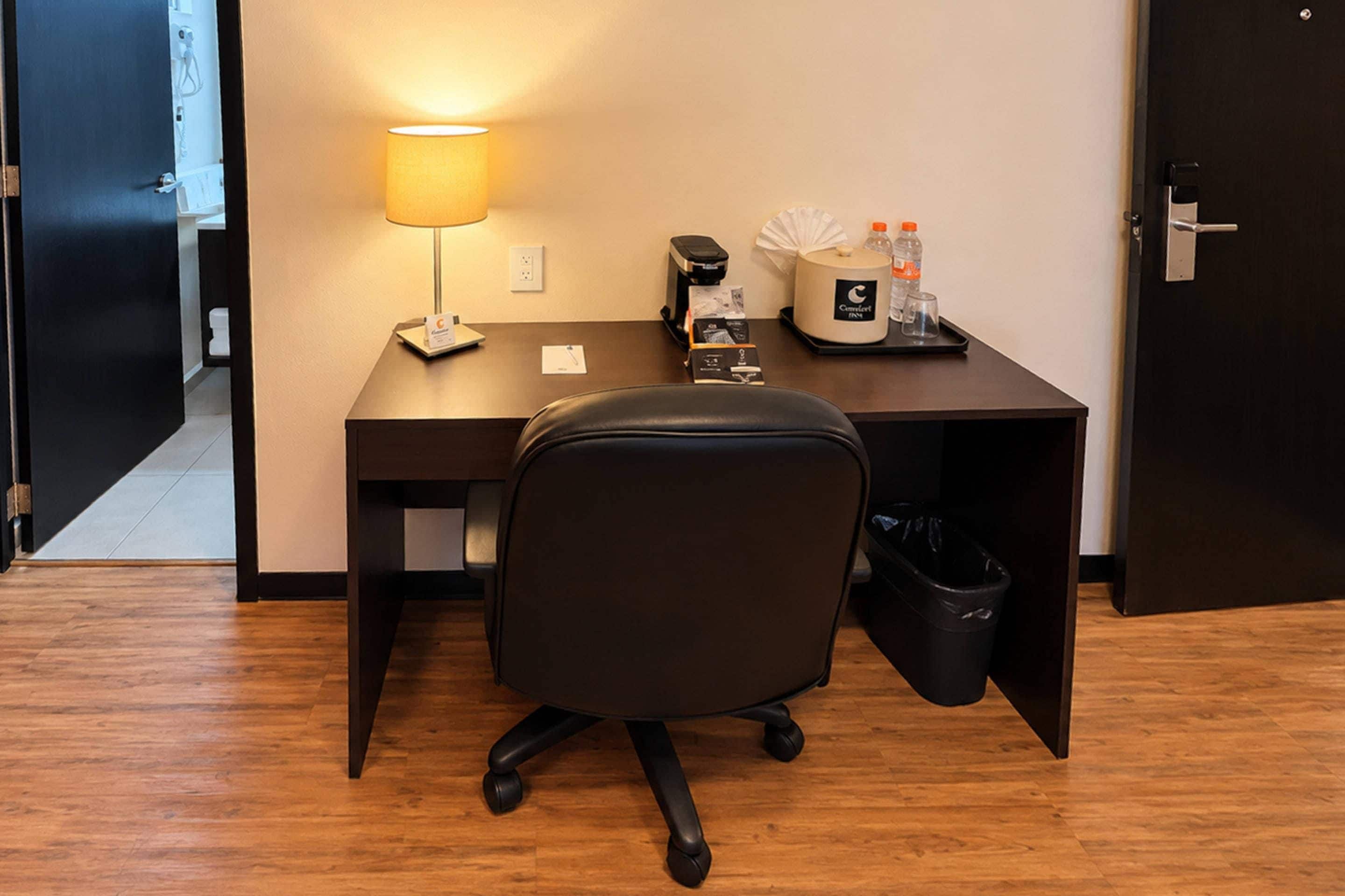 Habitación de hotel con escritorio, una silla, lámpara, cafetera, cubo de basura y minibar.