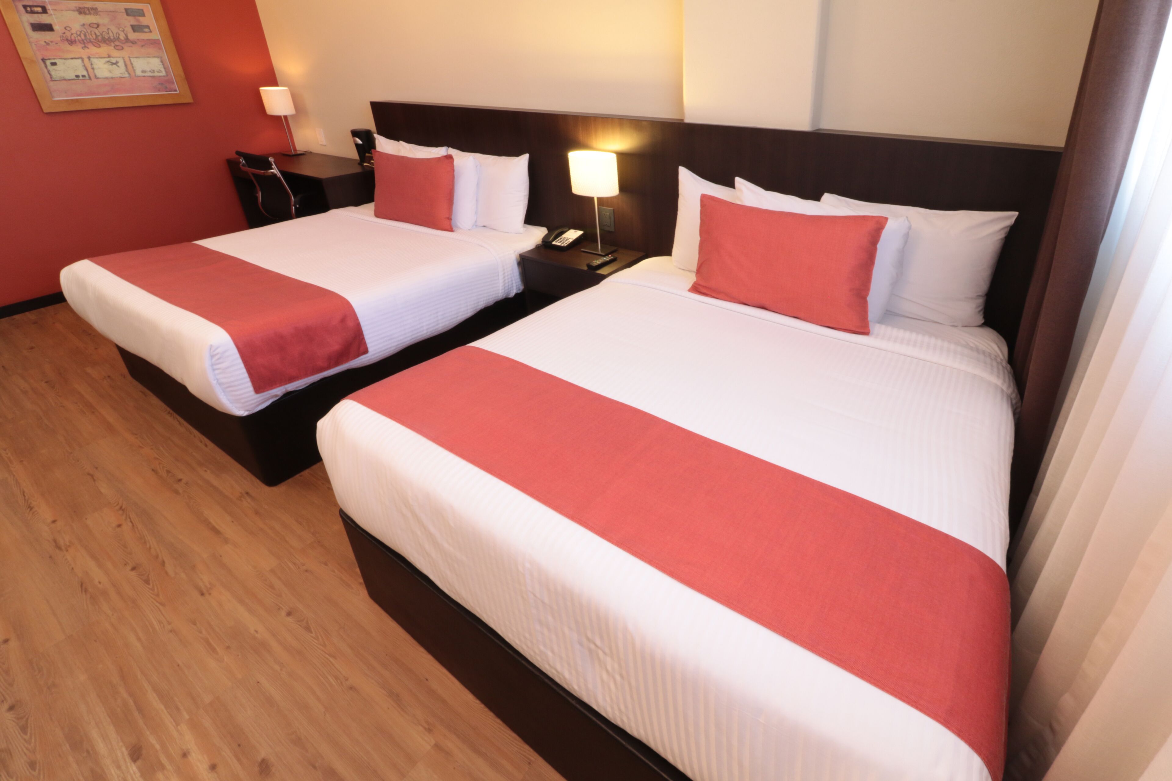 Habitación de hotel con dos camas, cada una con ropa de cama blanca y detalles rojos, una mesita de noche con lámpara y suelo de madera.